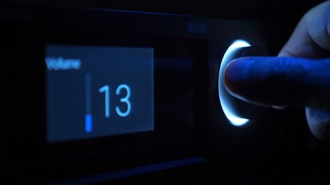 In the dark, the hand turns the volume control, illuminated by blue light on the front panel of the black stereo equipment. The display shows the music volume level. Closeup