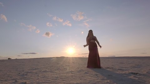 Beautiful young woman dancing oriental belly dance on sand against the sky at sunset.
