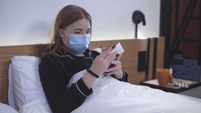 Ill Caucasian girl lying in bed in Covid-19 face mask and using video chat on smartphone. Portrait of infectioned teenager having symptoms of coronavirus viral infection. Pandemic concept.
