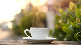 Close-up steaming hot coffee or tea cup on saucer, slow motion. Hot ceramic white coffee cup with smoke on old wooden table in nature background. Hot Coffee Drink Concept - Powered by Shutterstock - Get 15% off with code: PIKWIZARD15