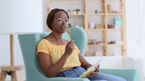 african woman with braids thinking and writing in notebook with pencil. Spbi creative female writer sit in living room. thoughtful young woman come up with ideas for business. concept list
