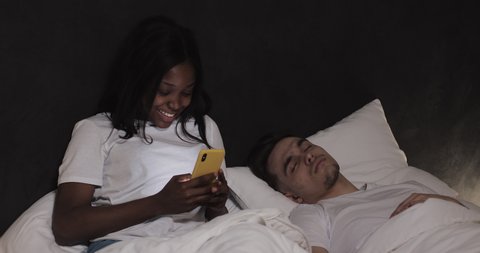 Happy multi-ethnic couple lying in bed at night under blanket. Young husband sleeping, while his wife handsome texting and cheating on his online via mobile phone