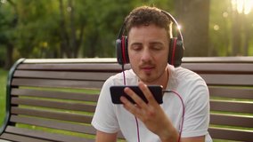 A young handsome guy in a white T-shirt and headphones sits on a bench in the park at sunset and listens to music on the phone or watches a video