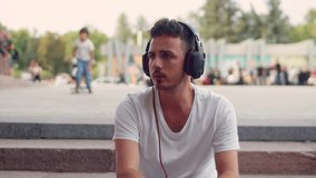 A young handsome man in a white T-shirt and headphones sits on the steps in the city and listens to music on the phone or watches a video
