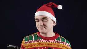 A young handsome man shows emotions - he is talking by video link with someone, he is wearing a red New Year's sweater with New Year's drawings and a red New Year's hat.