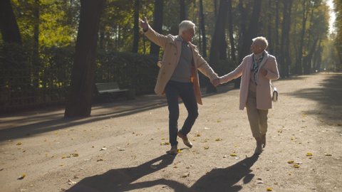 Happy old stylish couple dancing in fall park enjoying date. Senior man flirting with elderly woman walking in sunny city park. Active retirement and Autumn vibes concept