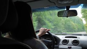 Young woman with long loose hair drives new car holding black leather steering wheel at designed dashboard along road past green trees cabin view
