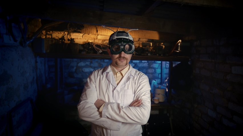 Funny professor in stupid glasses and a helmet. A scientist stands with folded hands in a dark garage. Lockdown. Home office. Teleworking concept.  Video meeting. Remote.