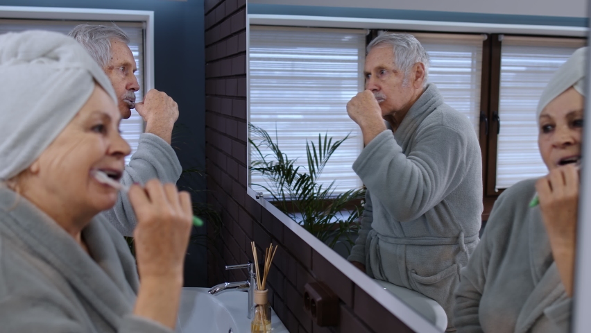 Cheerful old senior couple grandparents man and woman brushing teeth and looking into a mirror. Elderly grandmother and grandfather doing morning hygiene at luxury bathroom at home. Slow motion