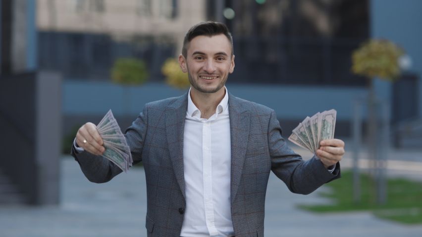 Amazed happy excited businessman with money - U.S. currency dollars banknotes. Man shows money and celebrating success, victory while looking to camera. Outdoors. Royalty-Free Stock Footage #1062630049