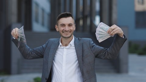 Amazed happy excited businessman with money - U.S. currency dollars banknotes. Man shows money and celebrating success, victory while looking to camera. Outdoors.
