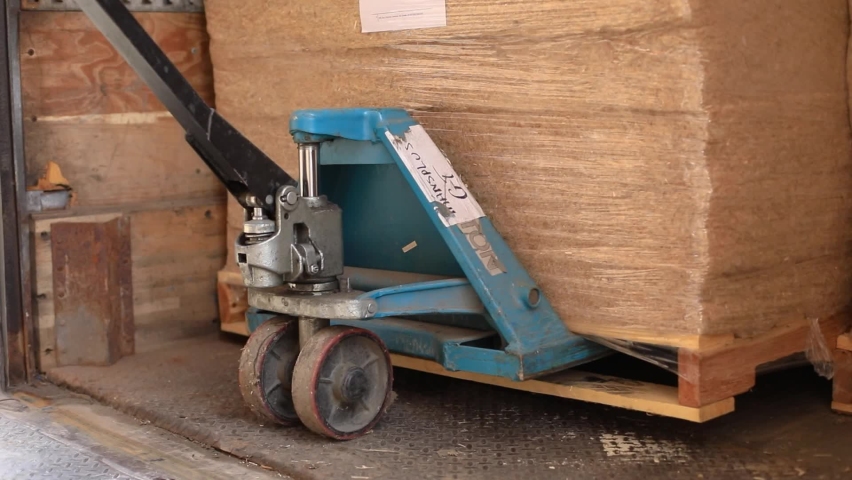 View from behing of carrier in orange safety jacket downloading from the back of a truck with a forklift pallets filled with sheets of hemp wool.