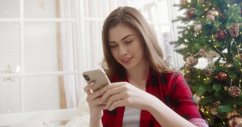 Close up portrait of joyful young beautiful Caucasian woman in room with christmas tree tapping and texting on smartphone wishing congratulation on Christmas Eve. Holidays concept