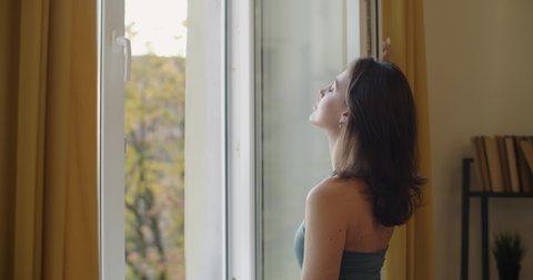 Side view of elegant woman opening window to breath fresh air slow motion. Sensual woman in sport bra standing by window looking outdoors natural light. Freshness energy sportive fashion concept