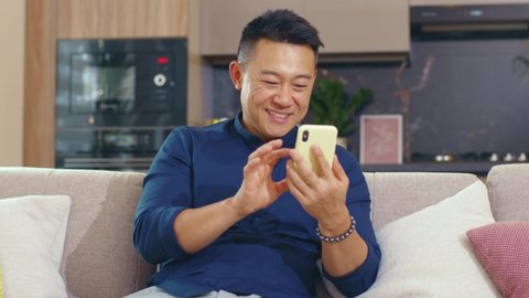 Smiling happy young asian man use phone sitting on sofa at home. Smartphone connection communication businessman chatting social media. Close up. Portrait. Slow motion