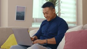Slow motion happy young asian man sitting on sofa use laptop computer work at home. Read good news. Network work smiling guy interior technology social media.