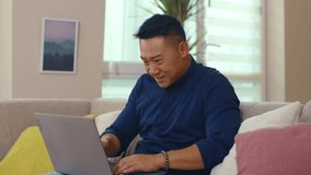 Happy young asian man sitting on sofa use laptop computer work at home. Read good news. Network work smiling guy interior technology social media. Slow motion