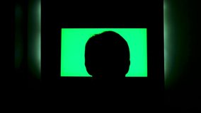 Rear view of a male head silhouette in front of tv with green screen. Concept. Man watching TV with chromakey that hanging on the wll at night at home.