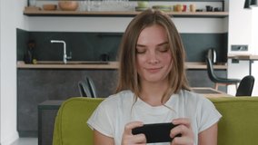 Smiling young woman playing cellphone app game at living room. Young caucasian female enjoying smartphone video games while sitting on sofa at living room.