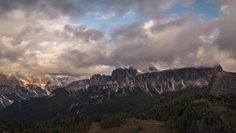 Clouds moving over the rocky mountain peaks of the dolomites in Italy, in the distance sunlight illuminates the landscape at sunset. You can see forests of pine trees with autumn colors in the valley.