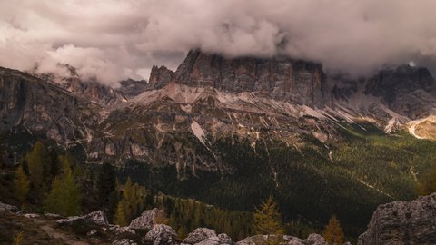 Motion time lapse of thick grey clouds moving over the rocky mountain peaks of the dolomites in Italy, in the valley below you can see forests of pine trees with beautiful autumn colors.