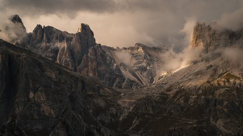 Clouds moving over the rocky mountain peaks of the dolomites in Italy with golden yellow sunlight illuminating the beautiful landscape. Light and shadow dance across the rock formations in the backgro
