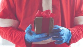 Santa Claus hold offers a gift box with ribbon bow in hands in protective gloves. Christmas shopping, New Year sales. Vibrant festive red color 4K video. Black Friday COVID-19 safe delivery concept.