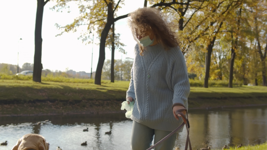 Young woman in safety mask picking up her pet poo with plastic bag outdoors. Female owner wearing protective mask walking with labrador and cleaning puke using plastic bag