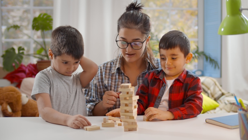 Happy young mother and two little children sitting at table and playing together. Nanny playing board game with preschool boys building tower with wooden bricks Royalty-Free Stock Footage #1062651478