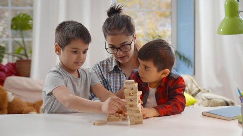 Happy young mother and two little children sitting at table and playing together. Nanny playing board game with preschool boys building tower with wooden bricks