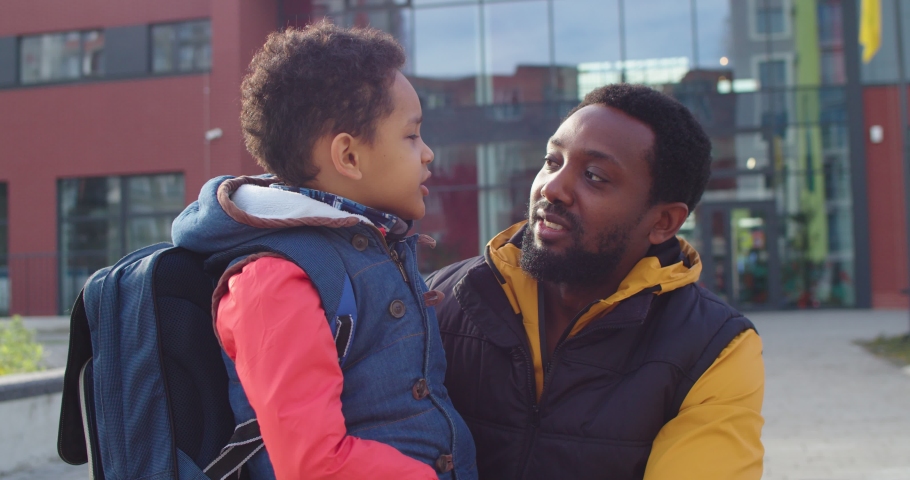 Close up portrait of joyful young man speaking with cute small son junior student outdoors in good mood on sunny morning. Happy African American little primary school pupil talking to father on street