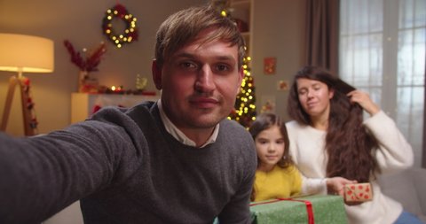 Close up of happy Caucasian handsome man with wife and little daughter waving hands while speaking on video call online in decorated room with glowing xmas tree on Christmas. New Year celebration
