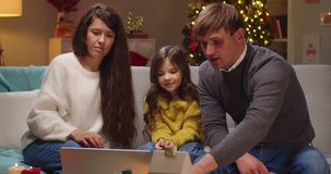 Portrait of Caucasian joyful family mom and dad with little girl together speaking on video call on laptop at decorated home spend holidays. New Year celebration. Holiday greetings