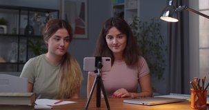 Caucasian happy beautiful young female twins waving hands and smiling while talking on online call on smartphone. Portrait of joyful sisters having video chat on cellphone at cozy home Leisure concept