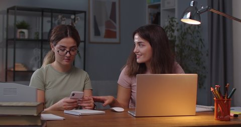 Portrait of Caucasian twin sisters sitting at desk in cozy room with laptop and smartphone. Female tapping on computer and talking with twin sister who is texting on cellphone. Indoors concept