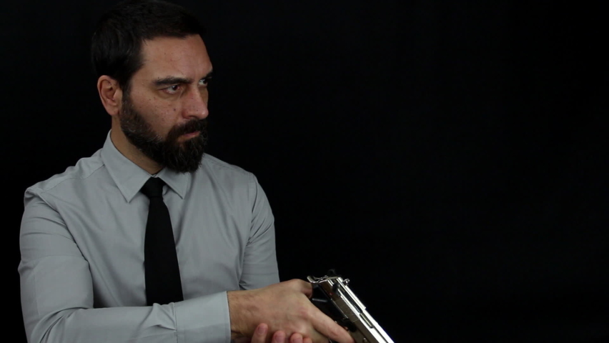 Handsome bearded man in shirt and tie on black background loads the shot in the barrel of his silver pistol and takes aim. Policeman dressed in civilian clothes