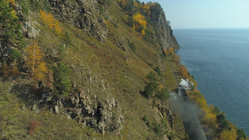 Aerial high Circum-Baikal Railway epic Transsib train, steam locomotive rides toneel railroad puffs of smoke. Autumn landscape Baikal lake. Rocky coastal nature. Russia Siberia travel landmark. 4k
