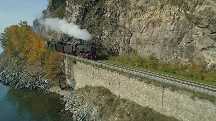 Flight backward Baikal lake train scenic steam locomotive blows white smoke rides along Circum Transsib railway along coast. Autumn unique natural landscape. Travel landmark. Sunny day. Aerial film