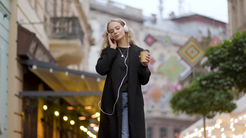 Slow motion of happy young woman in headphones walking outdoors in city street having fun alone. Joyful attractive blonde carefree woman listening to music and drink coffee