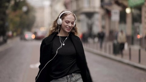 Slow motion of happy young woman in headphones dancing outdoors in city street having fun alone. Joyful attractive blonde carefree woman listening to music with smartphone