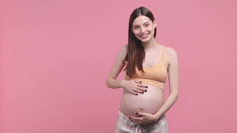 Happy pregnant woman future mom in basic top stroking keeping hands on belly stomach tummy with baby showing thumb up isolated on pink background studio. Maternity family pregnancy gynecology concept