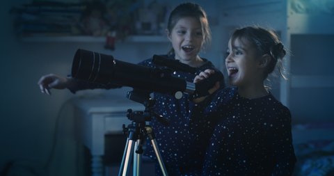 Cinematic night shot of two happy little girls sisters wearing starry pajamas are having fun to watching through telescope at falling stars in the dark sky. Concept: exploration, education, childhood