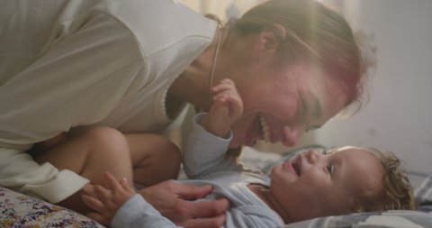 Cinematic close up shot of young happy mother is tickling and playing with her toddler baby boy in a nursery woke up in a morning. Concept of love, parenthood, childhood, life, maternity, motherhood.