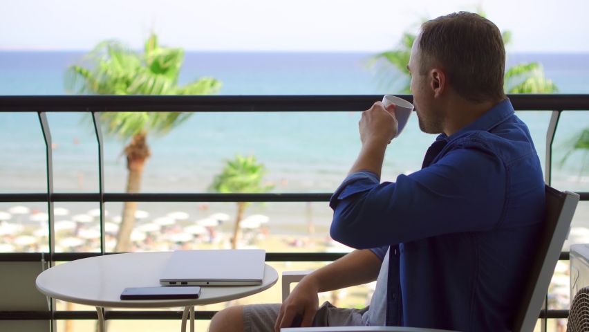Businessman sitting on balcony working on laptop at tropical beach resort. Young professional male freelancer type on computer and drink hot coffee or tea from white mug from remote home office | Shutterstock HD Video #1062664129