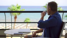 Businessman sitting on balcony working on laptop at tropical beach resort. Young professional male freelancer type on computer and drink hot coffee or tea from white mug from remote home office - Powered by Shutterstock - Get 15% off with code: PIKWIZARD15
