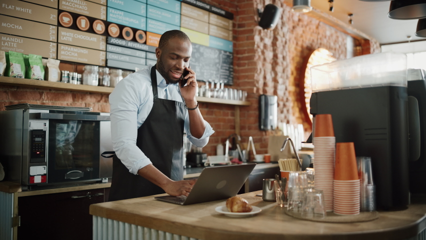Black African American Coffee Shop Employee Accepts a Pre-Order on a Mobile Phone Call and Writes it Down on Laptop Computer in a Cafe. Restaurant Manager Browsing Internet and Talking on Smartphone. Royalty-Free Stock Footage #1062664405