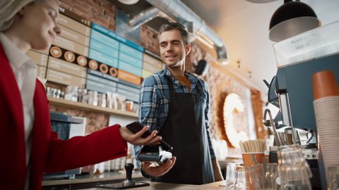 Female Customer Pays for Take Away Coffee with Contactless NFC Payment Technology on Smartphone to a Handsome Barista in Checkered Shirt in Cafe. Customer Uses Mobile to Pay Through Bank Terminal. Stock Video
