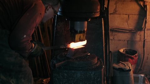 A mechanical hammer forges very hot metal. Man Blacksmith Forging Piece of Hot Iron Under Hydraulic Press at Workshop of Forge Factory. Metalworking. Blacksmithing Workshop.