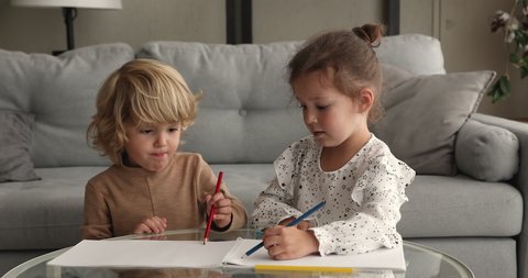 Little siblings sitting in living room at table drawing together pictures in sketchbook. Older friendly sister teaching to younger brother paint with pencils showing care. Friendship, hobby concept