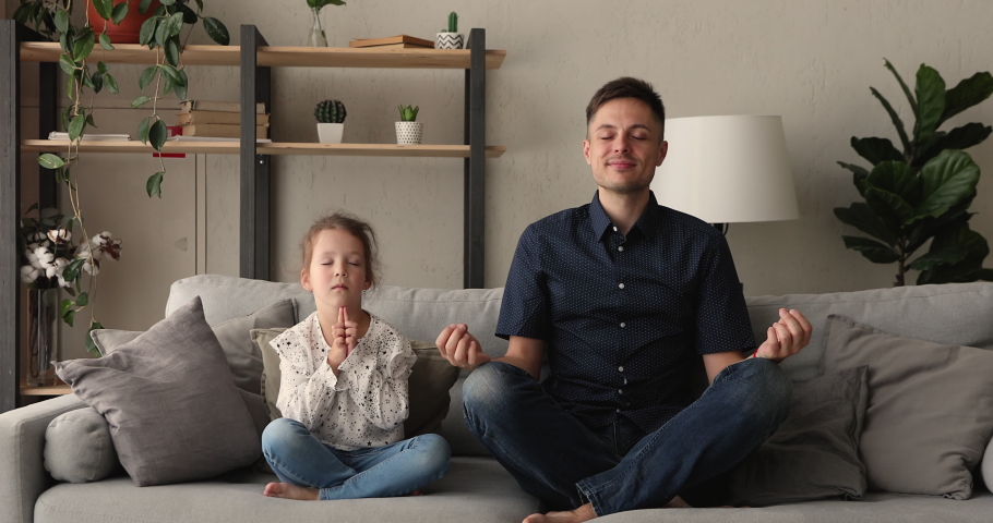 Tranquil father and little 5s daughter seated cross-legged on comfy couch closing eyes do together meditation asana, spiritual powerful practice. Teach kid manage emotions, healthy lifestyle concept Royalty-Free Stock Footage #1062671266